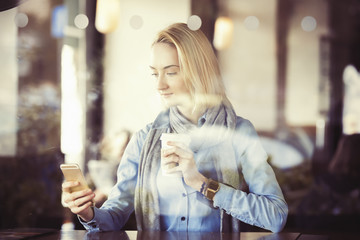 young woman is texting on her smartphone while drinking coffee, through glass