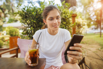 Cheerful beautiful woman is using an app in her smartphone device holding ice tea at the park