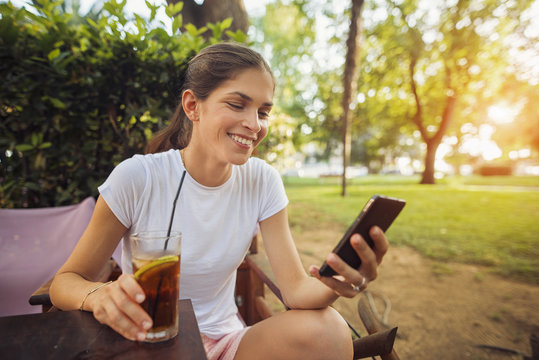 Cheerful beautiful woman is using an app in her smartphone device sending a text message holding and ice tea at the park