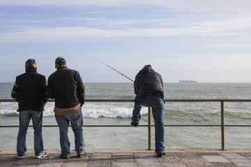 Drei Männer blicken auf das Meer hinaus, einer fischt
