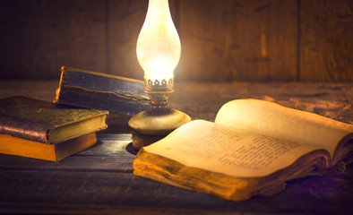 Old oil lamp and old books in darkness. Vintage kerosene lantern and open old book with blank pages on wooden table