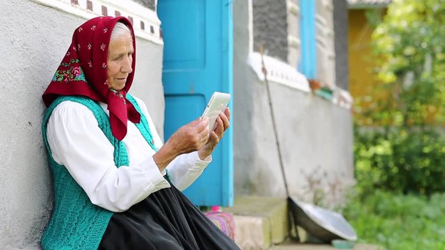 Old woman sits on bench and communicates via tablet computer. Senior woman sits on bench near his house and speaks via tablet pc in skype. Ukrainian old woman with tablet pc communicates through skype