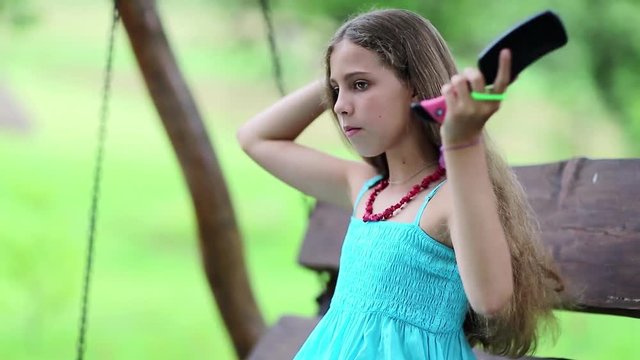 Attractive girl in blue sundress sits on the bench and combs his long hair. Beautiful girl combs his long hair. Girl with hairbrush sits on bench in garden