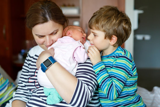 Little kid boy kissing newborn sister. Mother holding baby on arm.