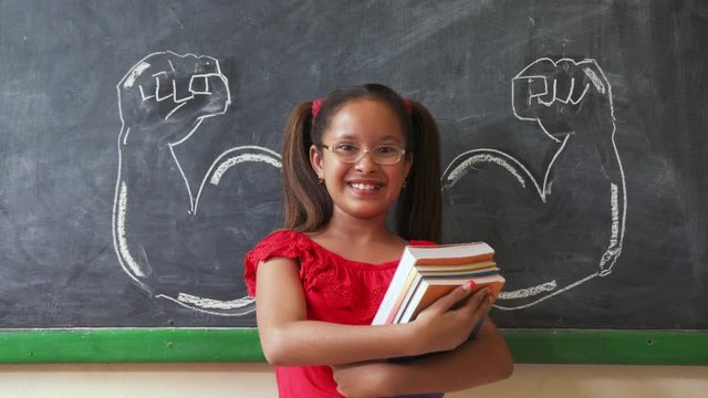 Concept on blackboard at school. Young people, student and pupil in classroom. Intelligent and successful hispanic girl in class. Portrait of female child smiling, looking at camera, holding books