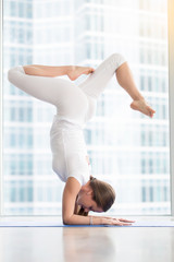 Fototapeta na wymiar Young woman practicing yoga, standing in variation of Pincha Mayurasana exercise with bent legs, handstand pose, working out, wearing white sportswear, indoor full length, floor window with city view