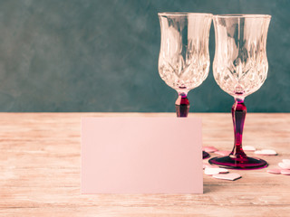 Two stemmed champagne glasses with pink hearts blank paper note card to fill with your text on wooden textured background. Valentine's day wedding romantic date invitation. Toned