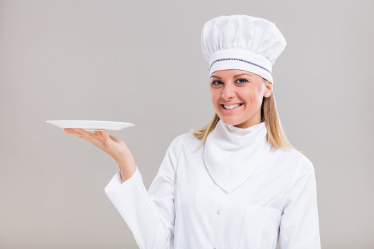 Beautiful Female Chef Is Holding Plate On Gray Background.
