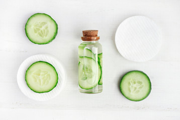 Liquid skincare cosmetic in bottle, green cucumber slice, morning facial fresh cleanser tonic, white wooden background top view.