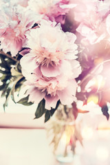 Lovely peonies bunch in glass vase on table with bokeh lighting. Romantic flowers bouquet, front view, pastel color