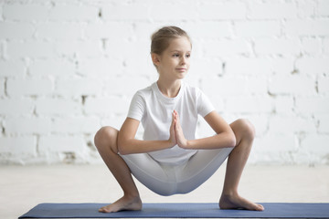 Girl child practicing yoga, stretching in malasana exercise, garland pose, working out wearing...