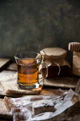 Glass cup with hot tea with jar of honey or jam, wood spoon, Spanish cookie polvoron on  vintage box, grey wall background,rustic vintage interior,cozy atmosphere