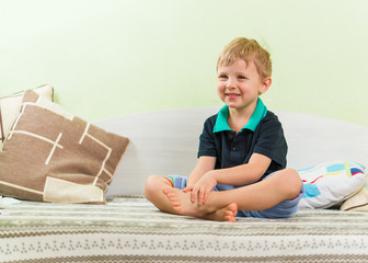 Cute skinny blond boy teeth smiling, sitting on a bed in the children's room, crossed legs and hands. Dressed in a casual black shirt and blue pants. Looking forward.