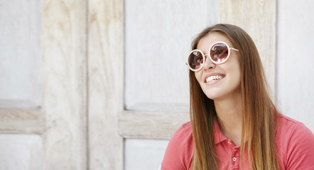 Fashionable teenage girl with charming smile relaxing indoors, wearing stylish round sunglasses. Attractive smiling young woman dressed in polo shirt at home. People, style and fashion concept