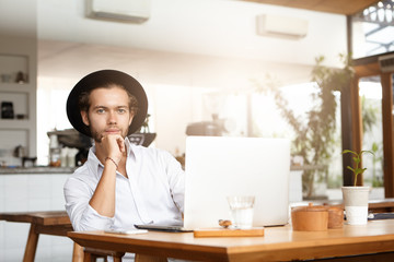 Stylish young Caucasian student in black hat having rest during work on diploma project, sitting at cafe table in front of open laptop computer, leaning on his elbow and looking at camera with smile