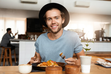 Fashionable male with beard appeasing hunger while dining alone at modern restaurant on sunny day, eating meal with knife and fork, sitting against blurred interior background and smiling happily