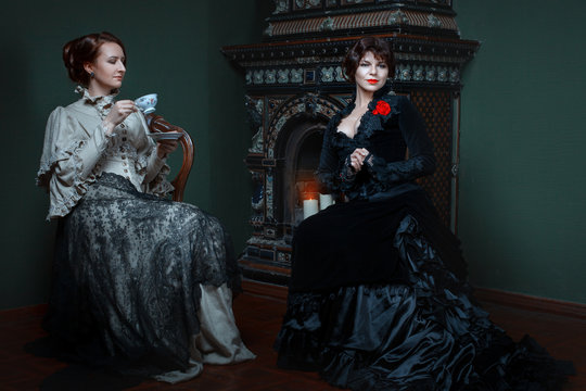 Retro woman drinking tea near the fireplace and lead the conversation, they are dressed in lavish dresses.
