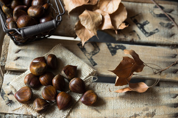 Fresh chestnuts in wire basket scattered on burlap cloth, dry brown leaves,on vintage wood box, autumn, fall, harvest, cozy atmosphere,top view