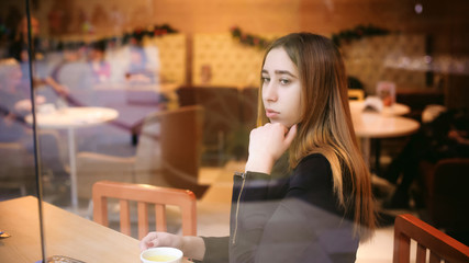 woman behind window in cafe. young girl in black dress sits at table, holding  beverage cup