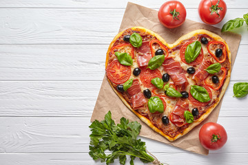 Heart shaped pizza love concept Valentine's Day symbol romantic dinner Italian food. Prosciutto, olives, tomatoes, parsley, basil and mozzarella cheese meal served on white wooden table background