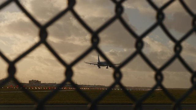 Plane landing on illuminated runway in early evening, seen through fence.