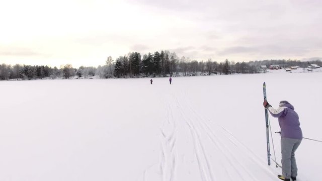Mature woman cleaning her ski while lagging behind. Daughter and granddaughter running towards to the lake shore. Russia
