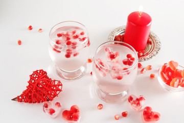 Romantic still life. Two glasses with alcoholic cocktail and ice, decorated with pomegranate, red candle, handmade heart, jelly candies, confetti. Saint valentine's day celebration.