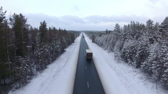 Camera back flying along winter road in evergreen woods of Karelia. Trucks driving on highway. The Kola route to the Murmansk; north of Russia
