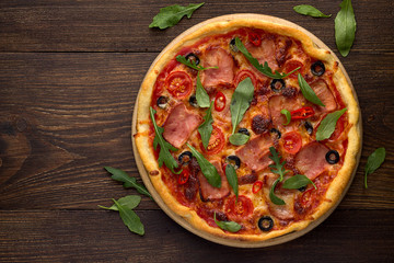 Italian pizza with ham, tomatoes, olives, cheese and scattered arugula on rustic wooden background.