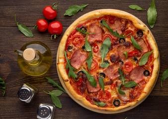 Homemade pizza with ham, tomatoes, olives and arugula on dark wooden table.