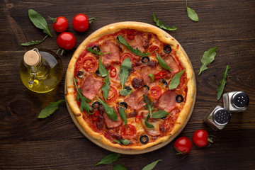 Traditional italian pizza with ham, cheese, tomatoes and olives on rustic wooden background.
