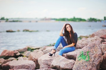 Beautiful pensive long-haired brunette woman in a black t-shirt, stylish jeans and shoes with high heels sitting on the rocks by the river