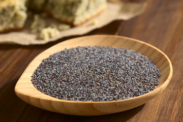 Rohe Mohnsamen auf kleinem Holzteller, mit Mohnkuchen im Hintergrund, fotografiert mit natürlichem Licht (Selektiver Fokus, Fokus ein Drittel in die Mohnsamen)