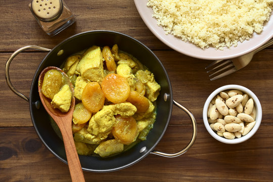 Chicken and dried apricot stew, seasoned with turmeric, cinnamon and honey, accompanied by couscous and roasted almonds, photographed overhead on dark wood with natural light