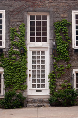 Fototapeta na wymiar White front door and sash windows. Green vines climbing up stone wall. Stone front steps and sidewalk in front of quaint town house in Old Montreal, Montreal, Canada.