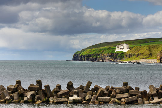 Dunbeath, Scotland - June 4, 2012: Brown, stone, protective pier in foreground, gray North sea and view of rocky green-covered cliffs with the white castle on top under big cloud sky.