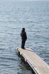 Man dressed in black with back to camera standing on a wooden dock jutting into Lake Baikal, Siberia. The water is a dark blue gray. 