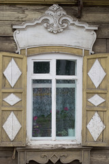 Window with tan shutters on an old wooden house in Siberia. Shutters have white wood diamonds. Window frames are carved.. Flowering plants are seen behind lace curtains.
