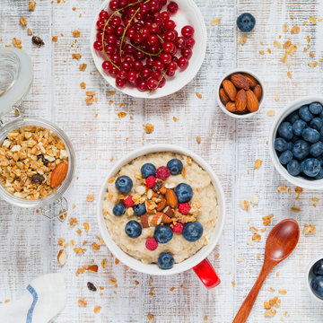 Healthy and nutritious breakfast: porridge oats with blueberries, raspberries and nuts in bowl with homemade granola and fresh berries served aside. Vintage white table background. Top view, square