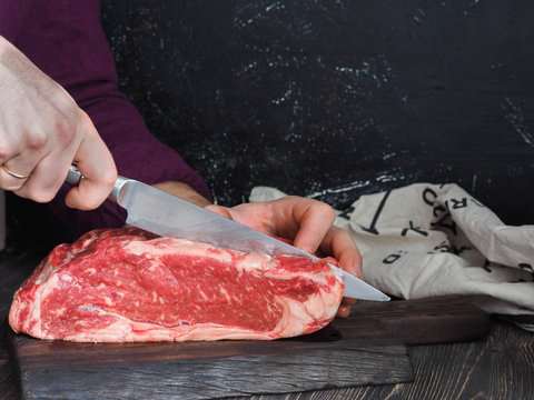 A piece of fresh beef on marble dark a wooden board. Male hand with a knife. Cutting the meat into steaks. Dark background.
