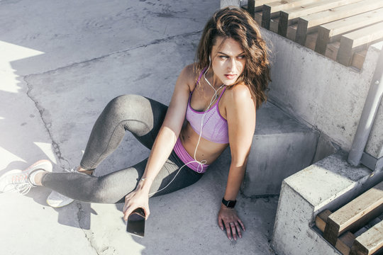 Summer sunny day. Young woman with brunette hair dressed in sport clothes, sitting outside on a concrete floor and listening to music on headphones. Girl resting after a workout, uses smartphone.