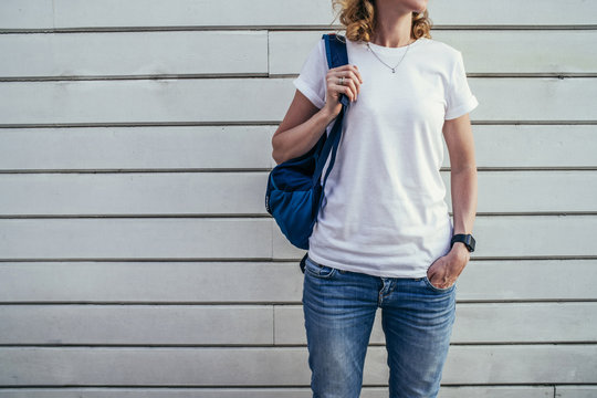 Close-up of young woman with backpack in white T-shirt and blue jeans standing outside with his hand in his pocket. In background white wall of wooden planks. On T-shirt space for text, advertising.