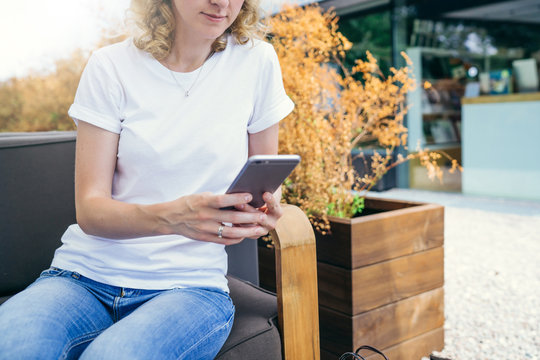 Summer sunny day.Front view,close-up of young woman in white T-shirt and blue jeans is sitting in street cafe and uses smartphone.In background plant in tub.On T-shirt with space for text,advertising.