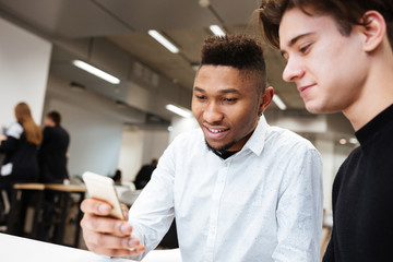 Young cheerful students sitting in library and using cellphone