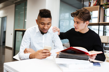 Two young students in library choose between book and cellphone.