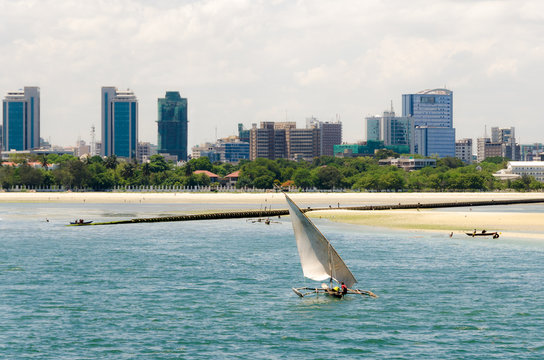 Coastline of Dar Es Salaam. A fishing boat is sailing along the beach towards the huge drainage pipe extending in to the ocean. The skyline with skyscrapers is in the background