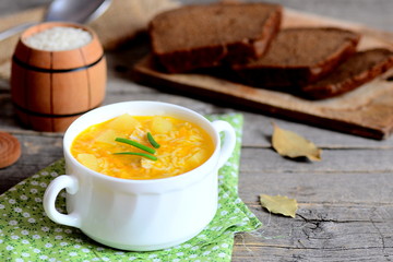 Delicious rice soup with chicken, potatoes, green onions and carrots in a bowl. Bread slices, green napkin, small decorative barrel with raw rice on wooden table. Quick and easy soup recipe. Closeup