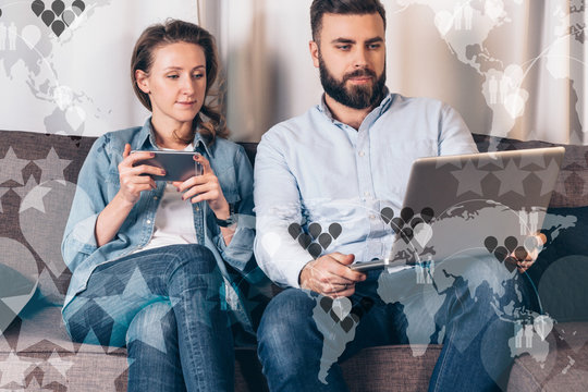 Bearded man and woman sitting indoor and choose on laptopt place for your holidays. Girl uses smartphone. In foreground semi-transparent virtual icons, image maps of the world and stars.