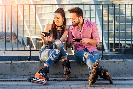 Smiling couple with cell phones. Two rollerbladers are sitting. Looking through our new photos.