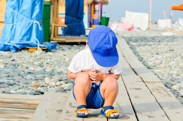 Boy in blue cap with sad expression. Resent and crouch. Frown baby boy on the stone beach in summer...